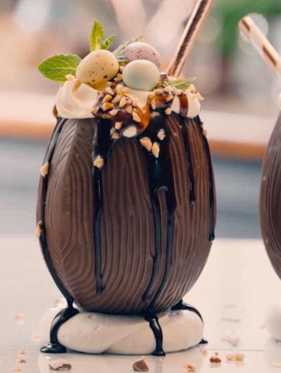 Deux cocktails d'œufs de Pâques en chocolat avec Baileys, crème fouettée, baies et mini œufs, avec une bouteille de Baileys en arrière-plan.