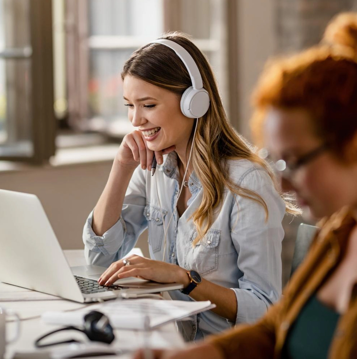 A smiling woman with headphones works on a laptop in a shared office