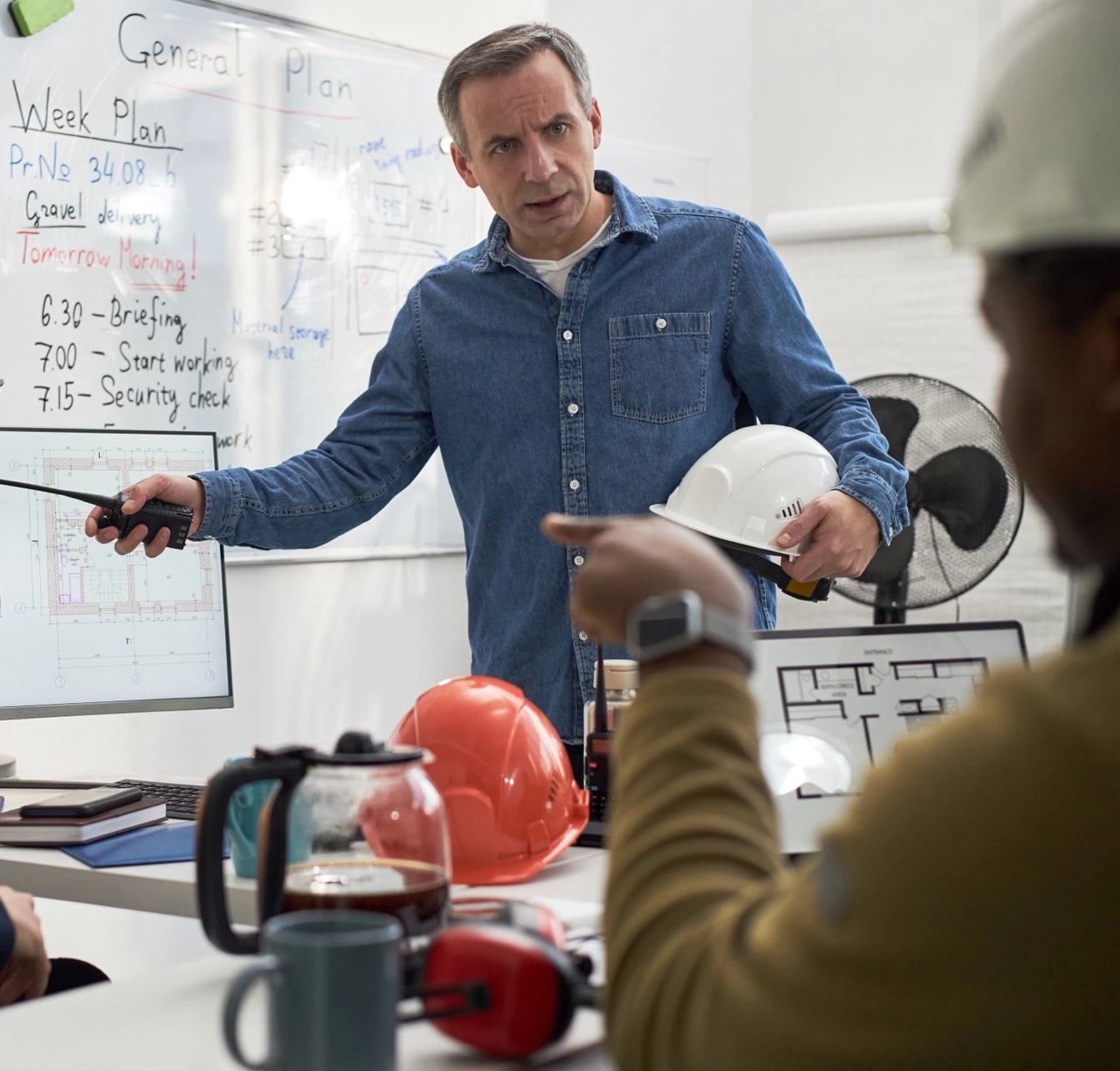 An engineer in a denim shirt holds a hard hat and talks through the week plan with his team in an office