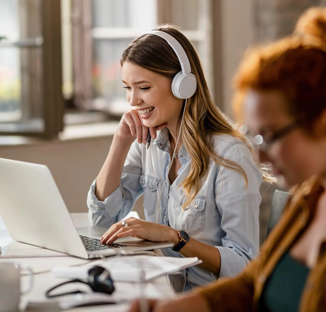 A smiling woman with headphones works on a laptop in a shared office