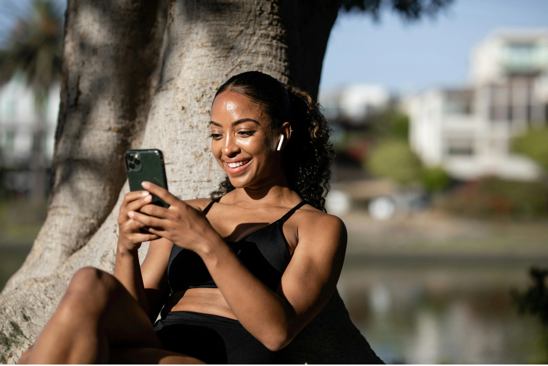 A woman sits outdoors in fitness attire smiling while using her iPhone