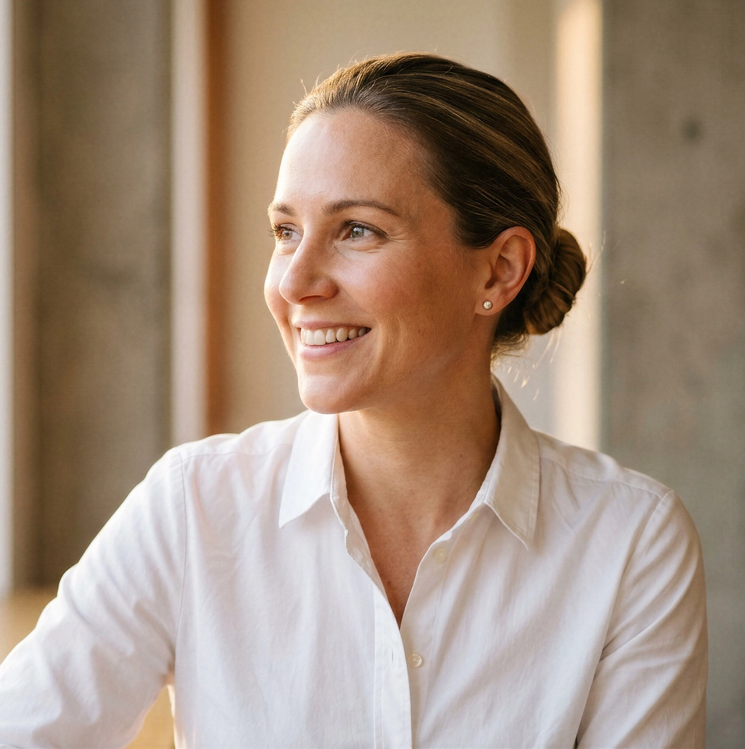 A smiling woman in a white shirt looks out of a window in a sunlit office