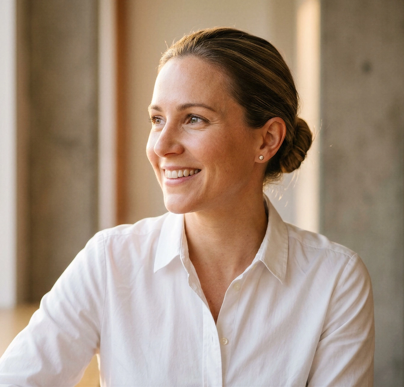 A smiling woman in a white shirt looks out of a window in a sunlit office