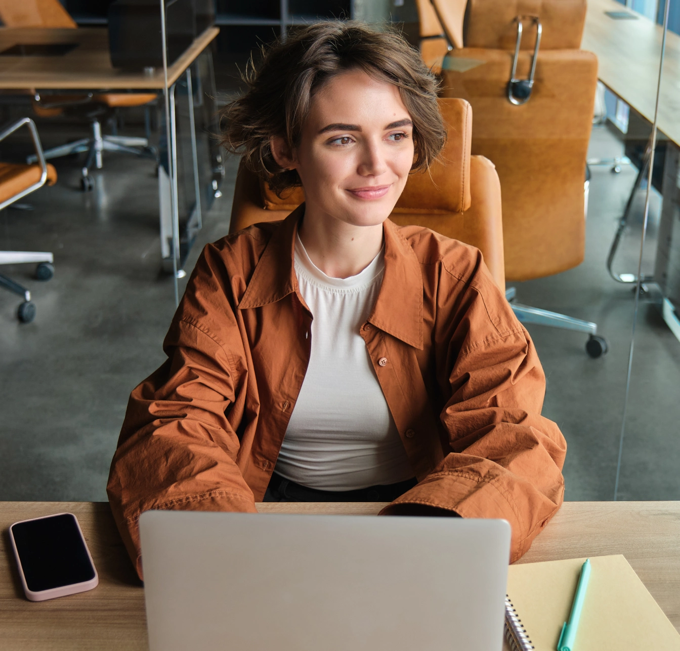 A smiling woman in a rust jacket sits at a laptop in a bright office