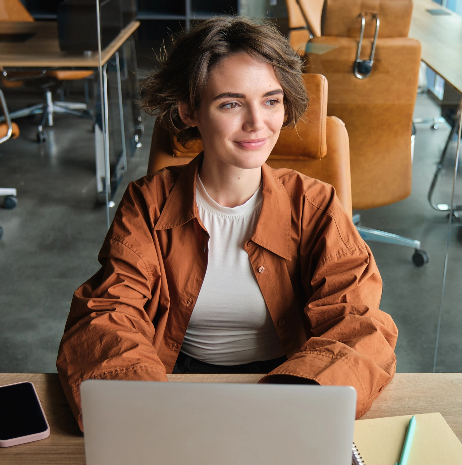 A smiling woman in a rust jacket sits at a laptop in a bright office