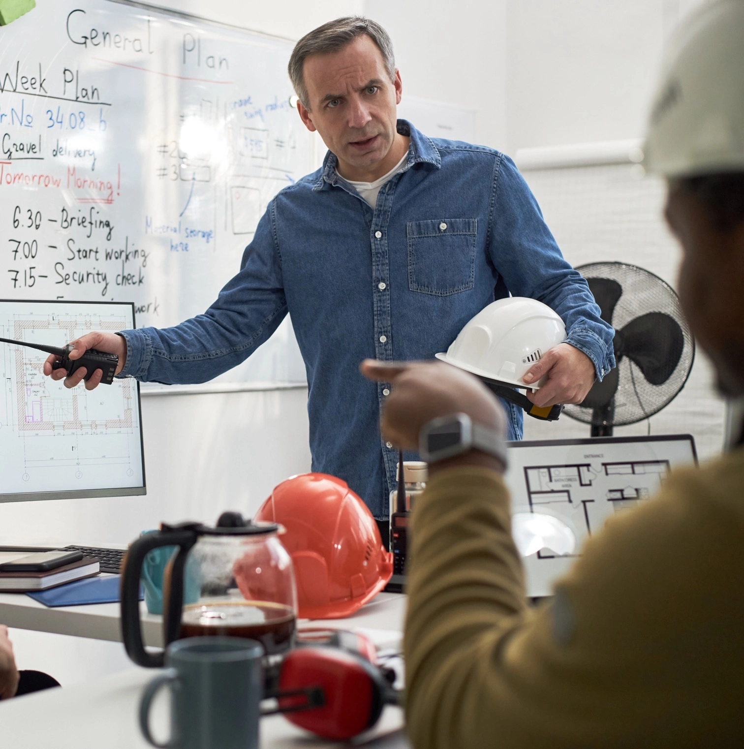 An engineer in a denim shirt holds a hard hat and talks through the week plan with his team in an office