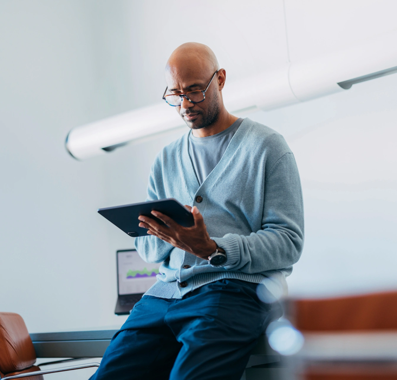 A man in glasses and a blue cardigan reads a tablet while sitting on a desk in a bright office