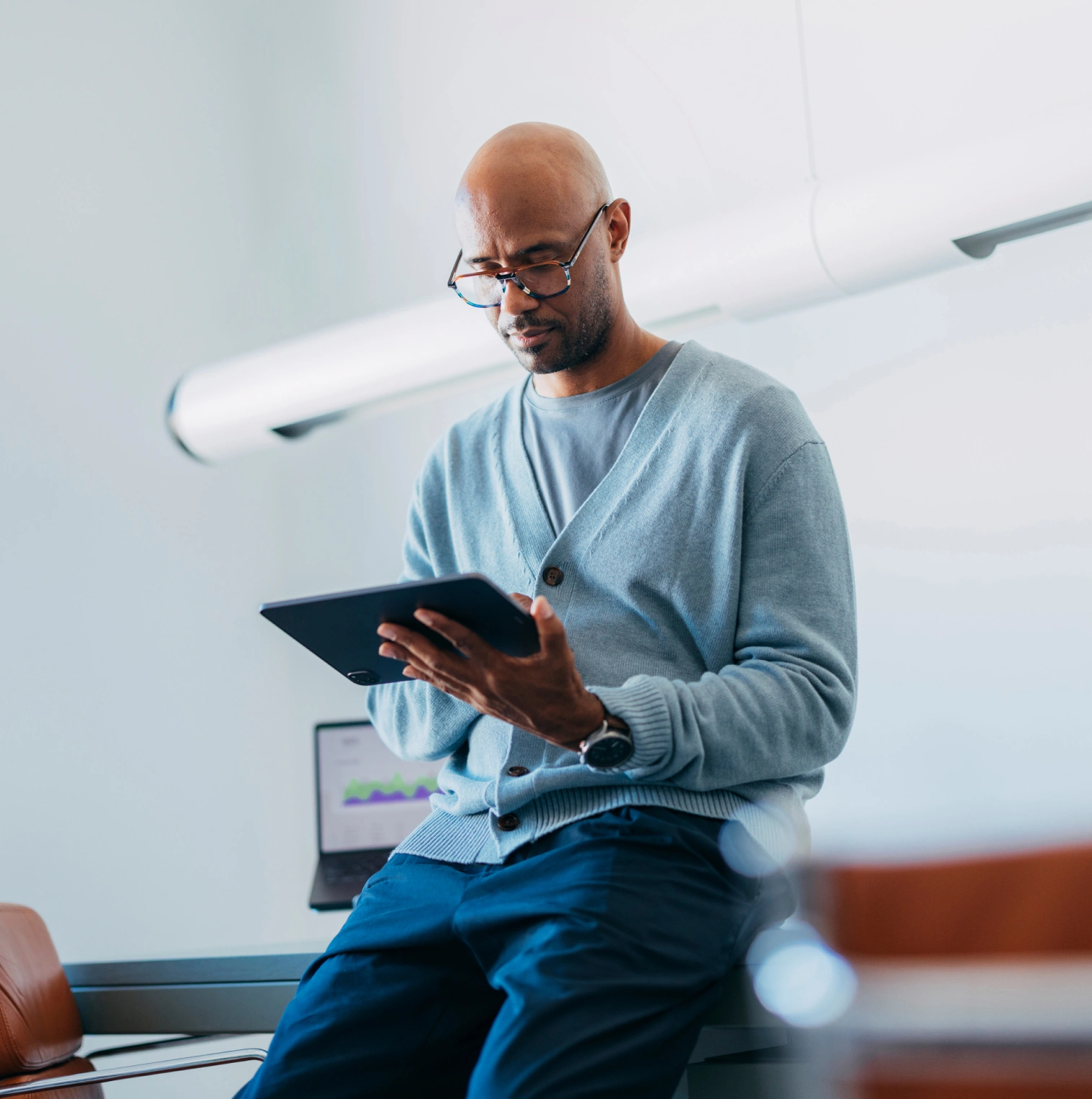 A man in glasses and a blue cardigan reads a tablet while sitting on a desk in a bright office