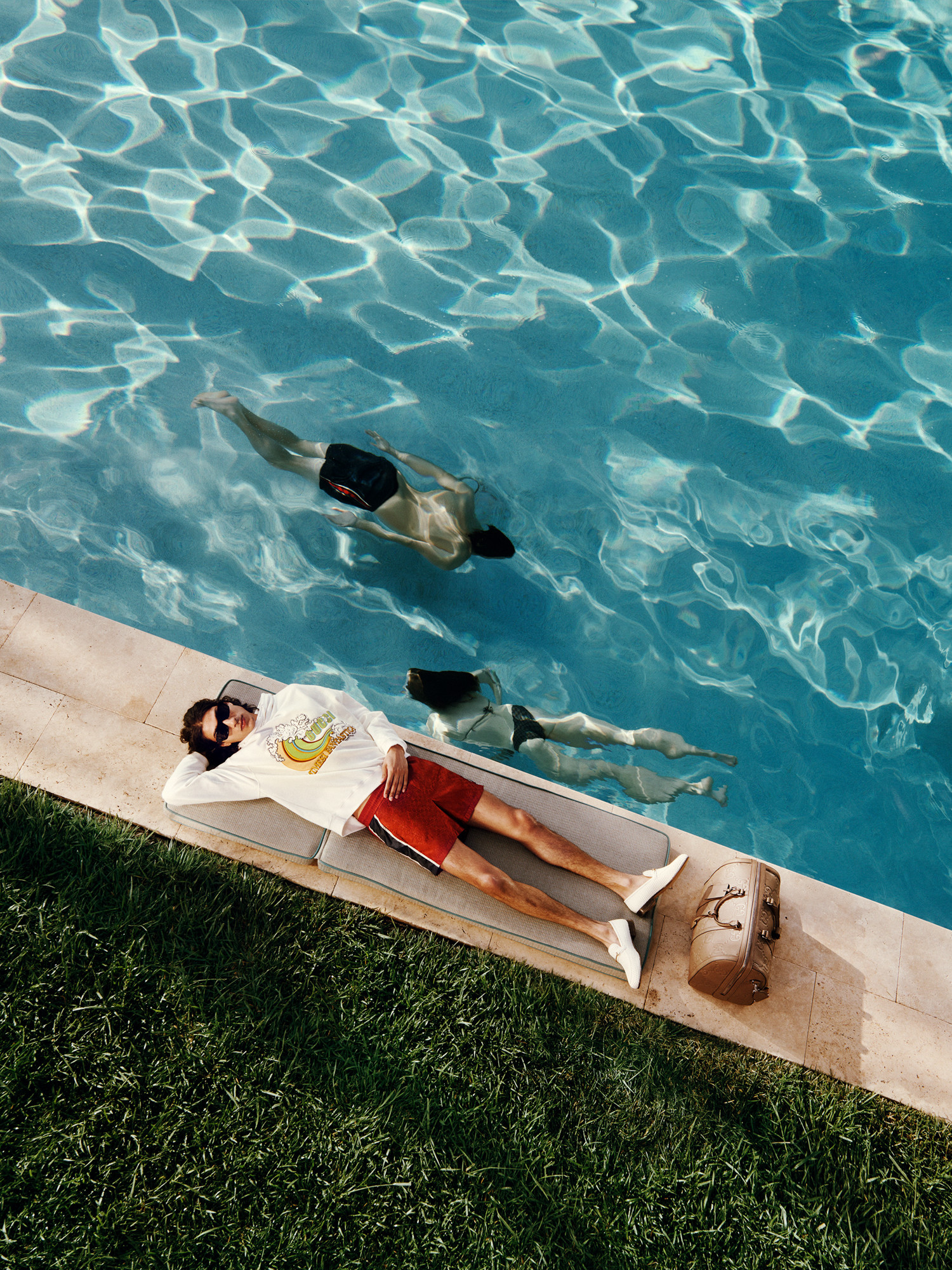 Un mannequin est allongé au bord d’une piscine. Il porte un sweat-shirt imprimé, un short rouge et des mocassins blancs. Un sac de voyage GG est posé à ses pieds.
