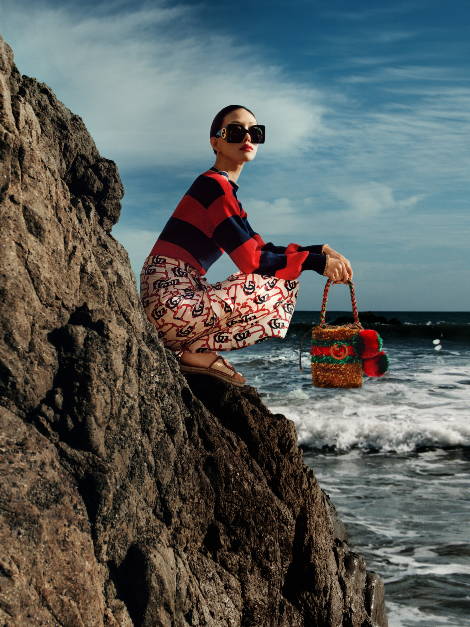 Model in a striped sweater and anchor print skirt on a sea cliff, holding a mini bag with Interlocking G logo, stripes and pompoms.