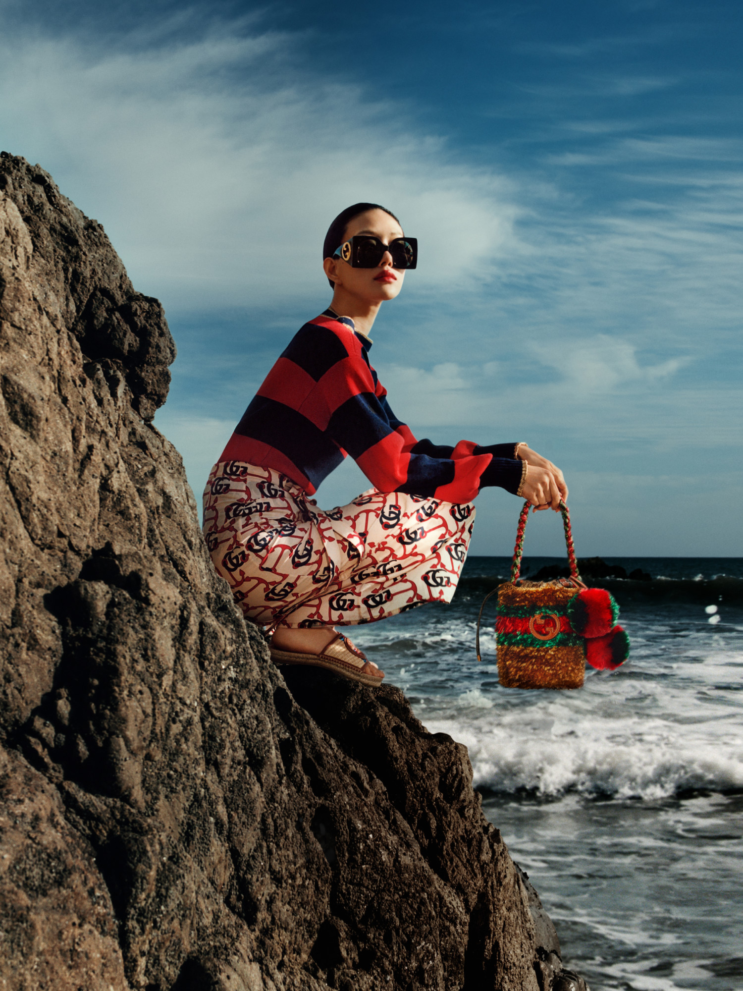 Ein Model mit einem gestreiften Pullover und Rock mit Anker-Print, das eine Mini-Tasche mit GG Logo, Streifen und Bommeln hält und an einer Klippe über dem Meer steht.
