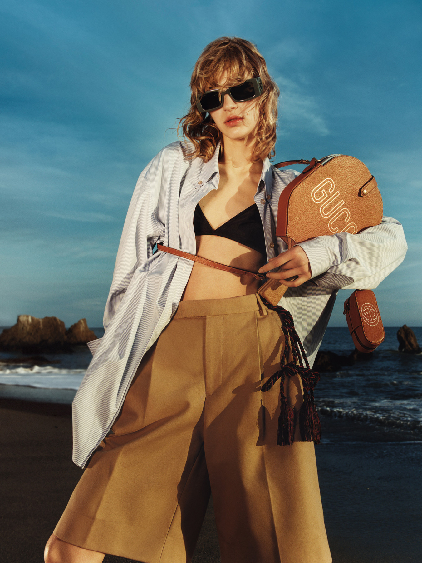 A model holds a Gucci logo beach racket, wearing a blue button-down shirt, black bikini and brown pants. She's at the beach.