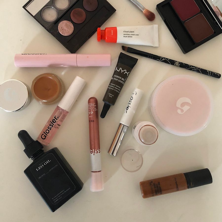 Glossier Lash Slick, Cloud Paint, Lidstar, Priming Moisturizer Rich, Boy Brow, Lip Gloss, and Stretch Concealer with other make up products on a table.