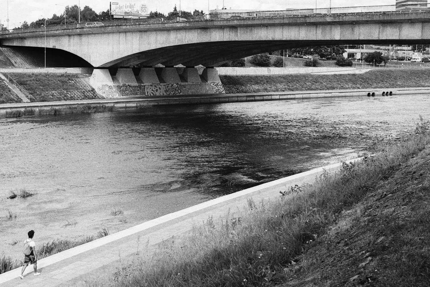 A man walking near the river, in front has a gigantic Vilnius bridge