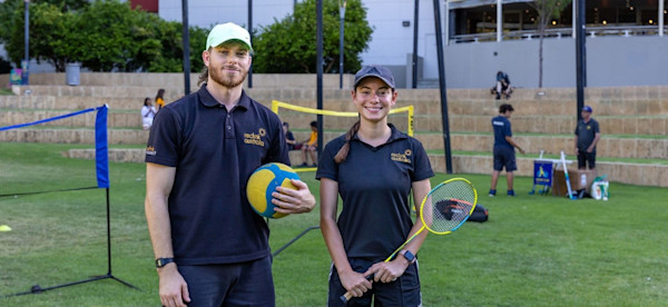 Two adults holding sporting equipment with young people and volleyball nets in the background