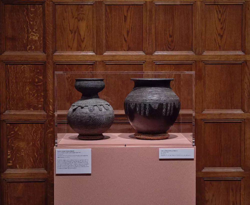 Two dark clay pots set in the same exhibition room against another wooden wall. On a pink plinth