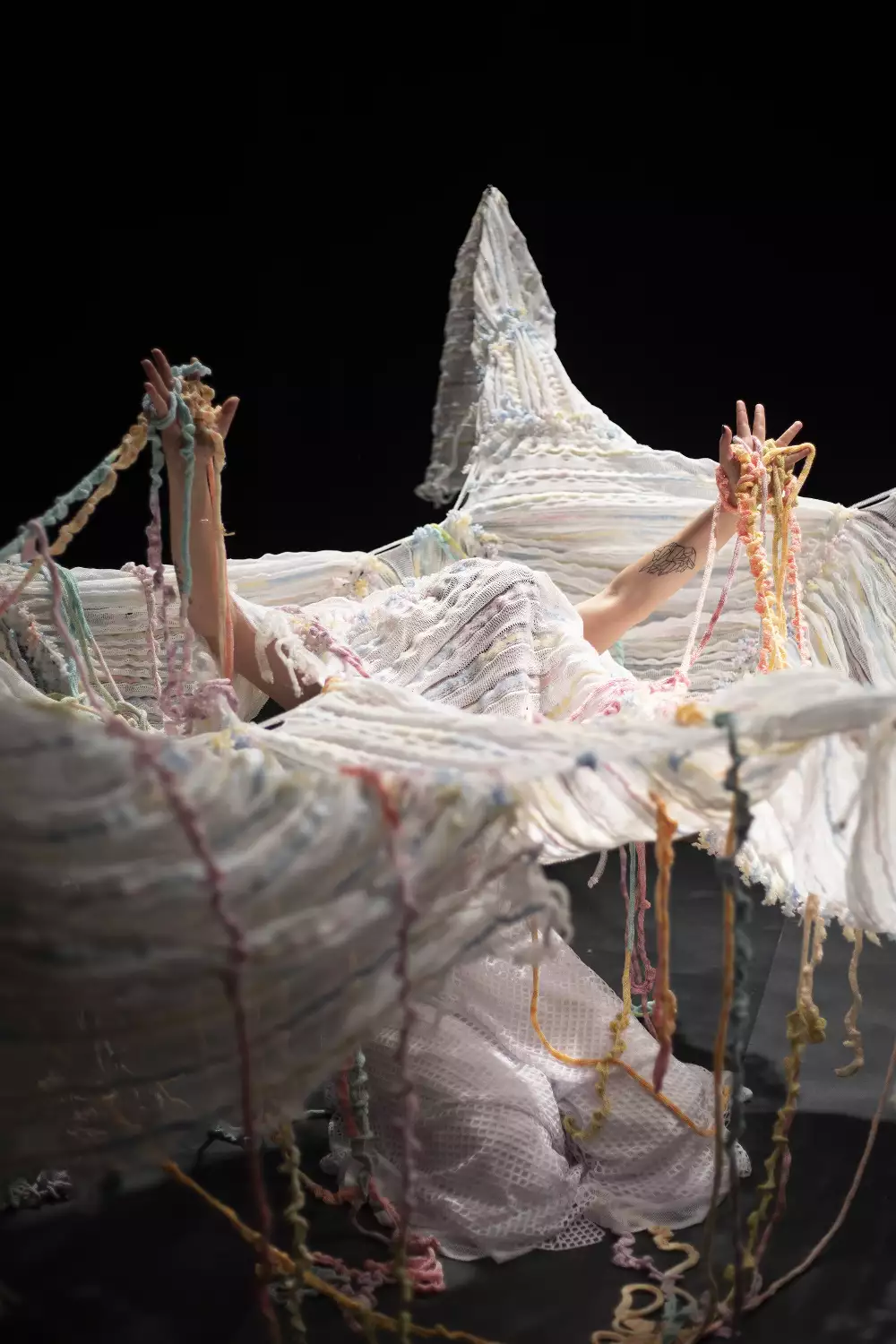 A woman tangled within a large knitted piece of yarn, performing in a dark room