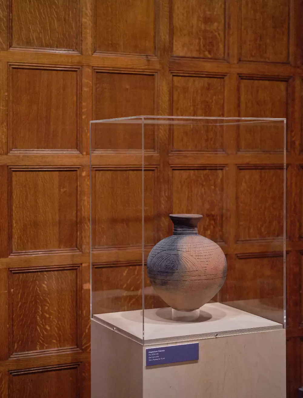 Image shows a clay, multi-tonal vase on a grey plinth inside a glass case. it is set against a wooden panelled wall