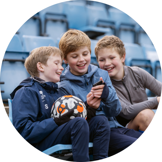 Bilde av tre unge fotballgutter som sitter på en tribune, smiler og ser på mobilen.