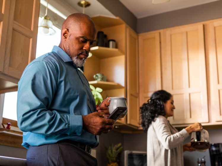 Man in kitchen holding coffee and looking at phone while woman pours coffee
