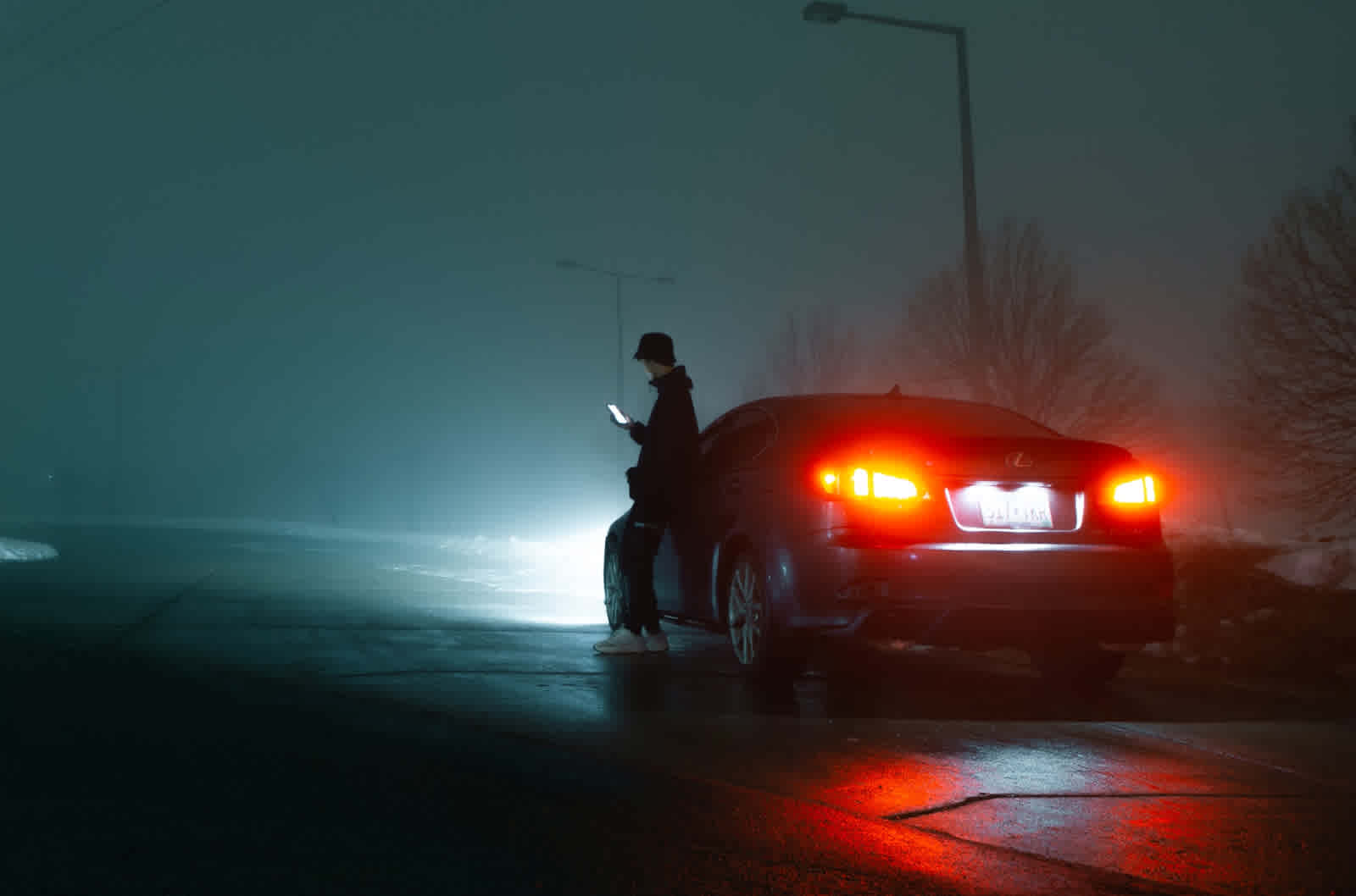 A person stands outside their car looking at their phone on a foggy night