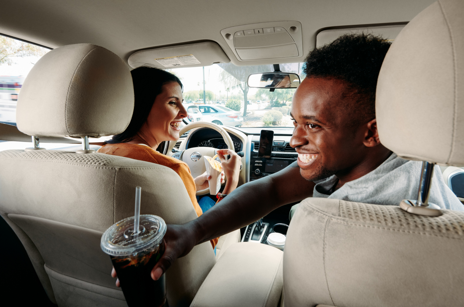 Two people sit in the front seats of a car, the passenger is turned around passing a drink to someone in the backseat