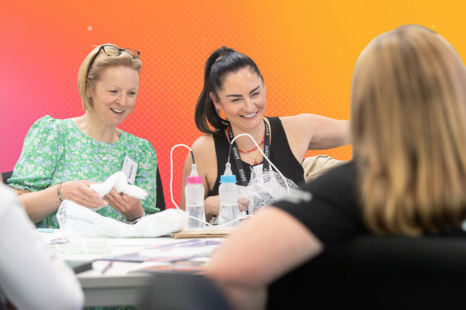 Two women sit at a group desk laughing whilst taking part in an experiment using wires and baby bottles. The image has been superimposed on a gradient background.