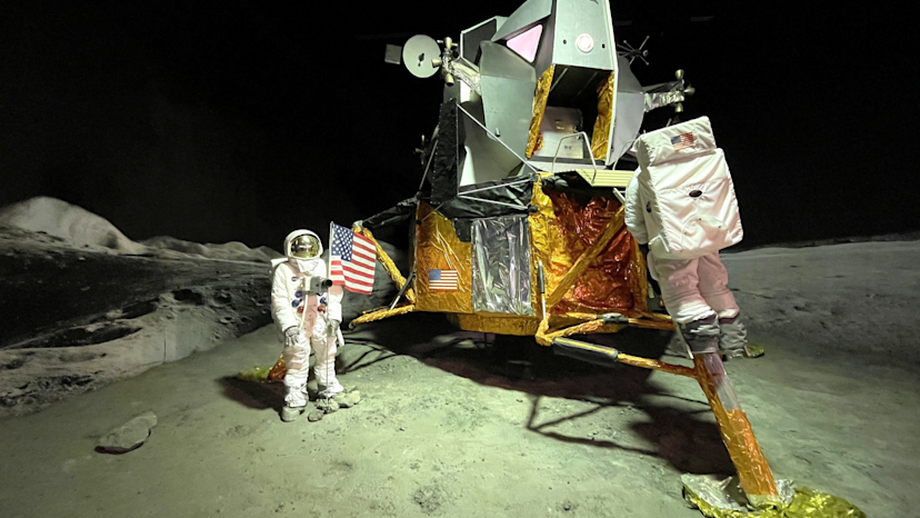 Image from the original moon landing of the lunar craft. An astronaut stands to the left with the american flag and an astronaut with his back to the camera is seen climbing up the craft.