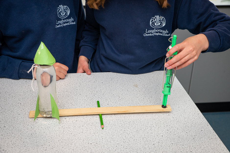 Two children measuring the movement of a small rocket, balanced upon a plank of wood being used as a lever.