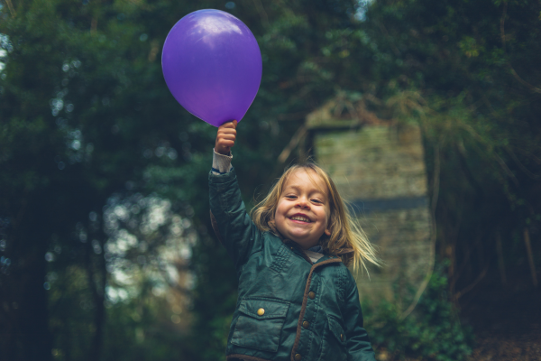 Nursery-aged child stands outside beaming with a coat on as they hold up a balloon in one hand. 