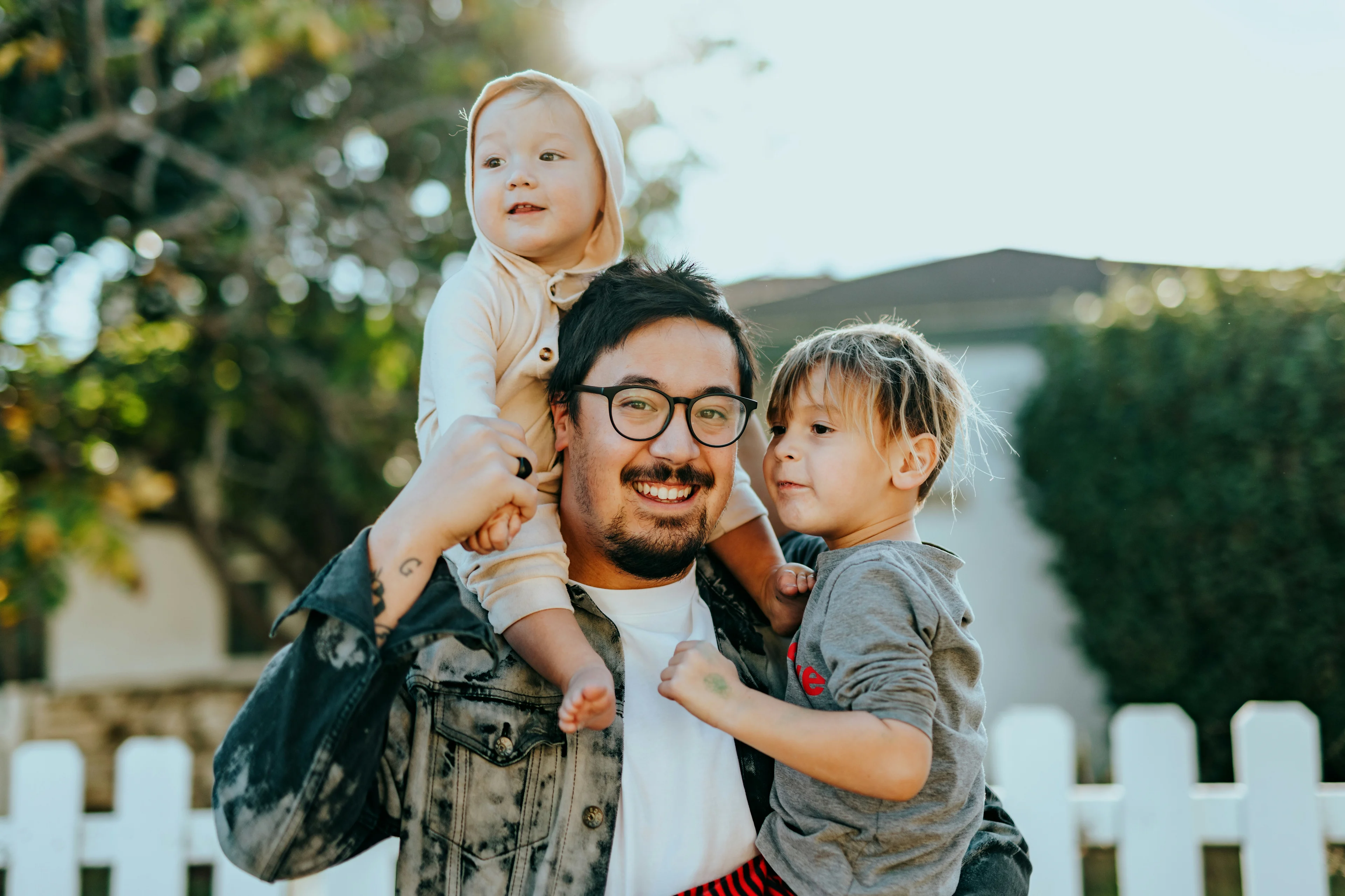 A smiling man holding an infant on his shoulders and a toddler in his arms