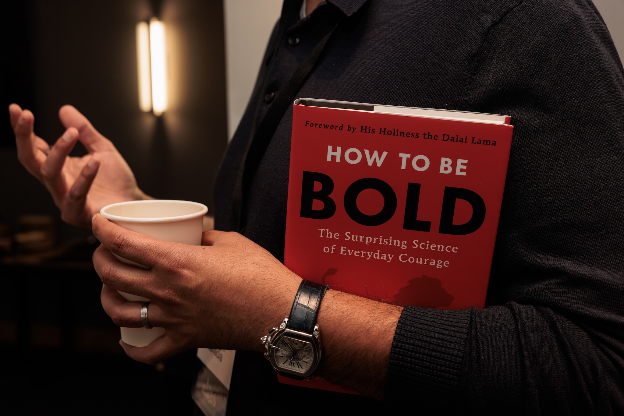 Book with the title "How to be bold" in the arms of someone holding a paper cup and wearing a watch.