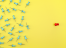 An assortment of blue thumbtacks scattered across a vibrant yellow background. Among them, a single red thumbtack stands out on the right side, pushed firmly into the surface.