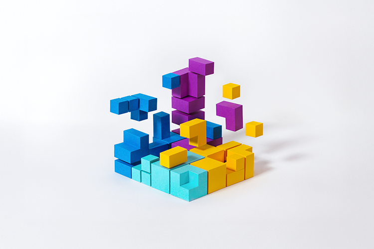 An abstract structure created by an arrangement of colorful, three-dimensional cubes in varying sizes and heights. The cubes are in shades of blue, yellow, purple, and teal. Some cubes are stacked, creating a layered appearance, while others appear to be floating above the structure.