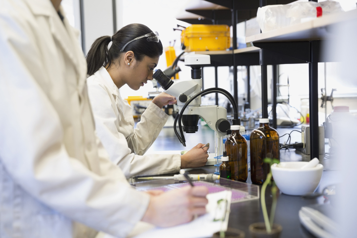 A person in a lab setting looks through a microscope and takes notes while another person in the foreground writes on a clipboard.