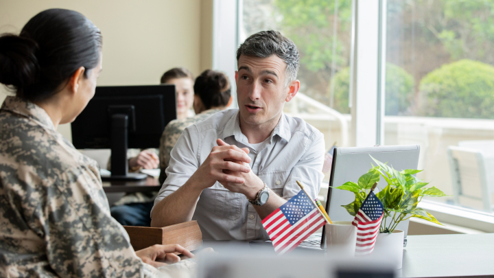 The image shows the back of an army soldier in uniform talking to someone sitting at a desk. Small American flags decorate the desk.
