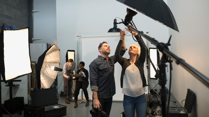 Two people in the foreground adjust a light on the set of a photo shoot with two people in the background.
