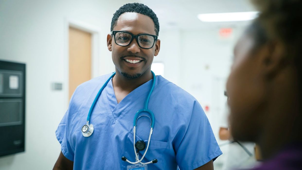 Ein medizinischer Mitarbeiter in blauen Kitteln mit einem Stethoskop um den Hals lächelt