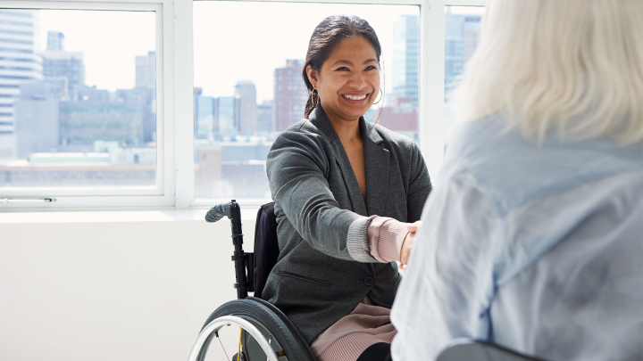 A person in a wheelchair wearing a low ponytail smiles shakes the hand of another person whose back is to the camera