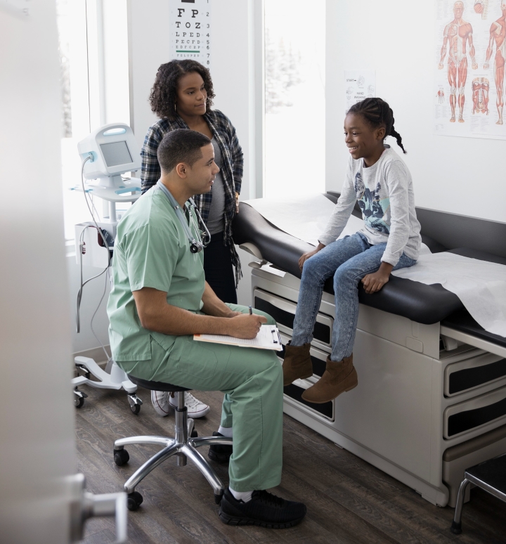 A health care professional sits on a stool while talking to a young patient sitting on a medical exam table as an adult looks on. The professional in light green scrubs has a stethoscope and a medical file.