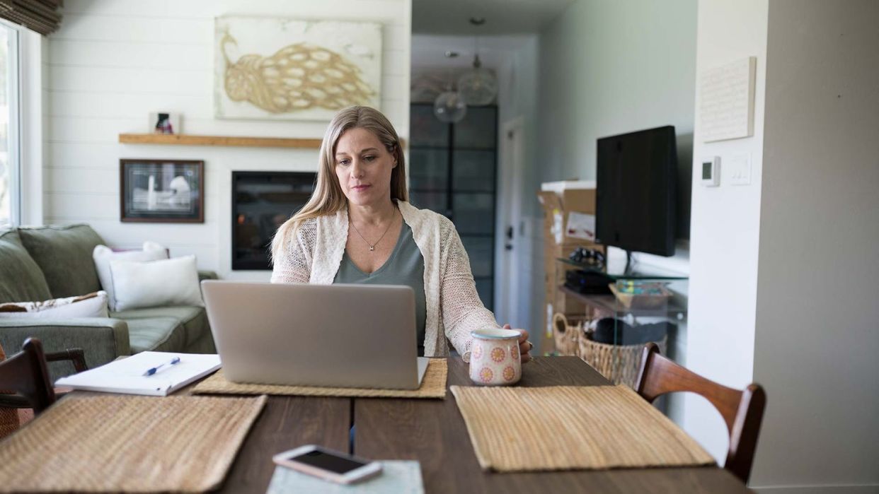 A person sits at a dining table facing their laptop while holding a mug.