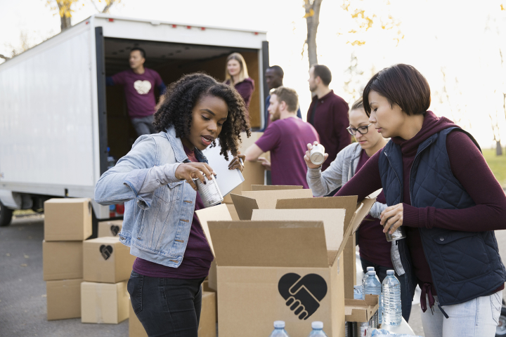 A person adds canned food to a box to be loaded on a truck. Several others are packing boxes and loading them onto a truck. They all wear matching T-shirts.