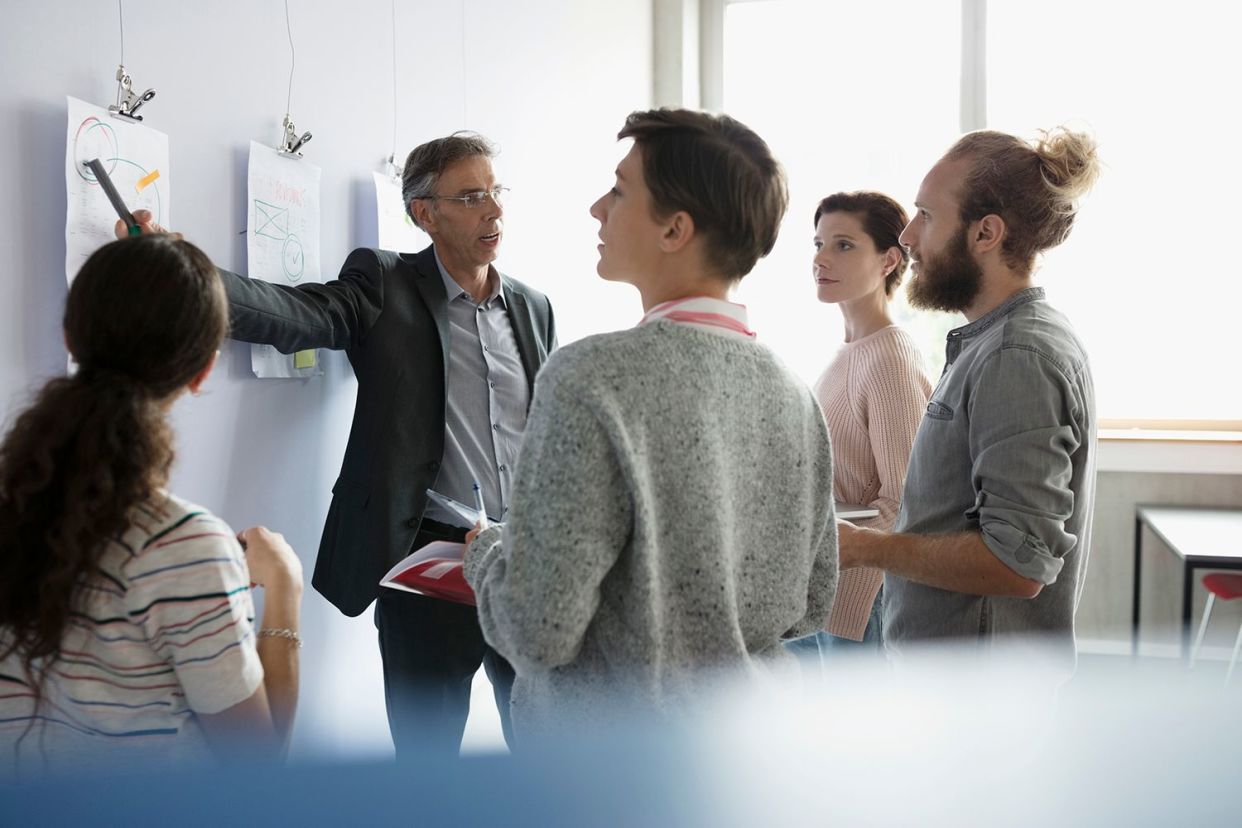 Eine Gruppe von vier Personen steht um eine Person herum, die auf ein Diagramm an der Wand zeigt.