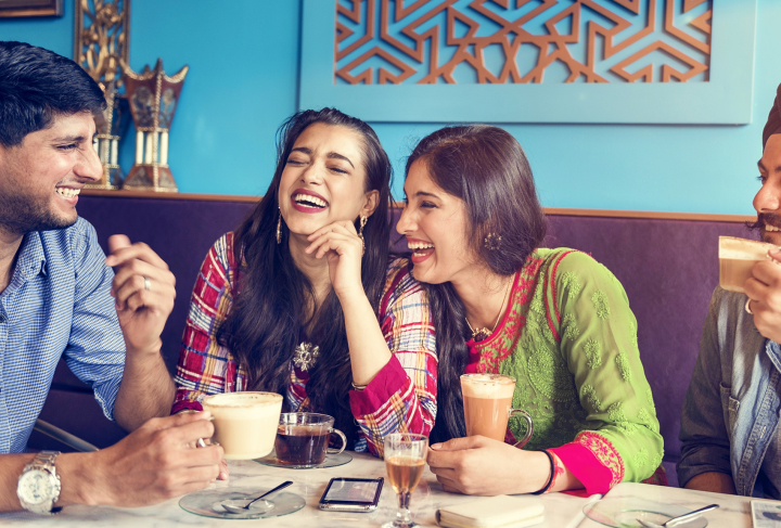 A group of friends sits around a table laughing and drinking coffee and tea.