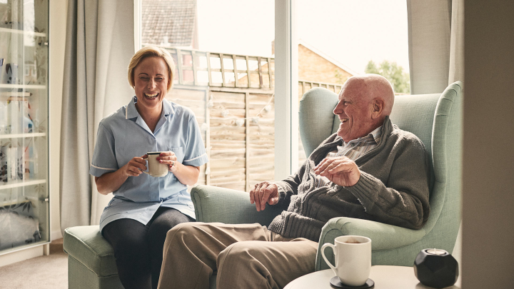 A home health professional or caretaker laughs with an older person sitting in a chair. The professional wears a uniform top and holds a cup of coffee or tea. Another cup is on a table next to the patient or client.