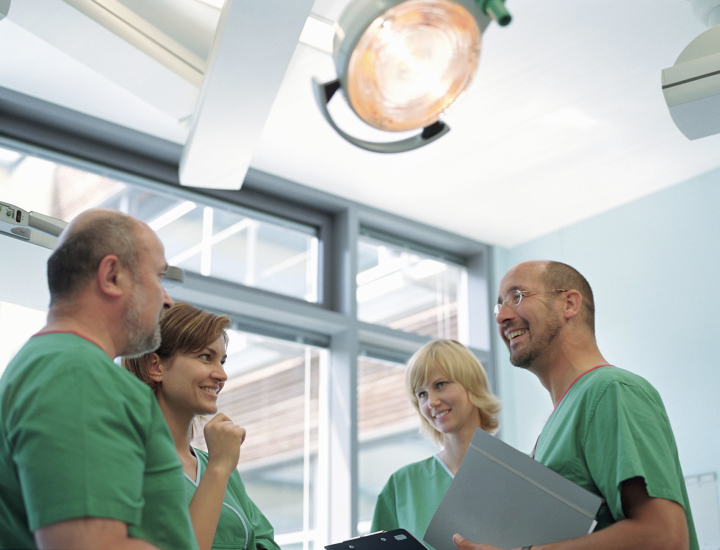 Four medical professionals in light green scrubs stand facing each other in a medical room. Two hold medical files. A large examination light hangs overhead.