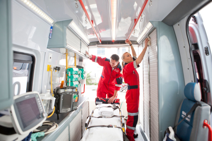 Two people in matching red uniforms wipe down cabinets in an emergency medical ambulance or transport. Medical equipment is on a nearby shelf. A gurney is in the center of the vehicle's aisle, and the back and side doors are open. 