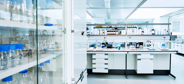 A workbench in a stark white clinical setting with bright overhead lights shows testing equipment. Supplies or testing materials are displayed in a glass cabinet.