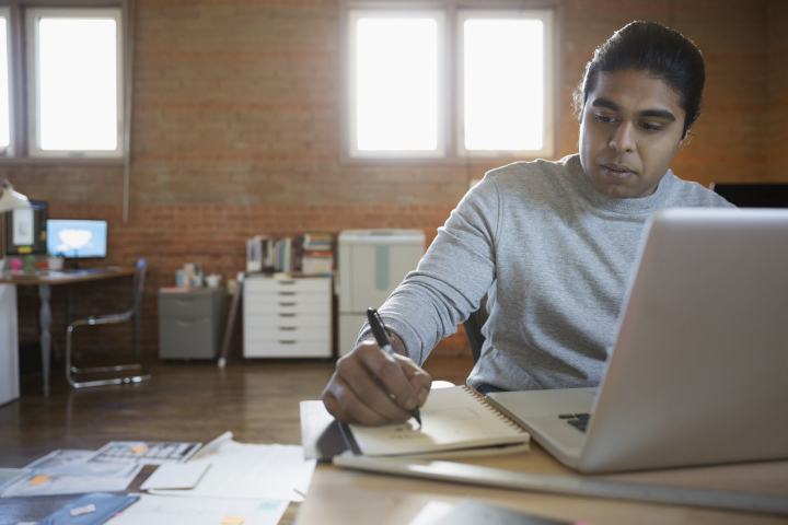 A person seated at a desk uses a laptop and writes in a notebook.