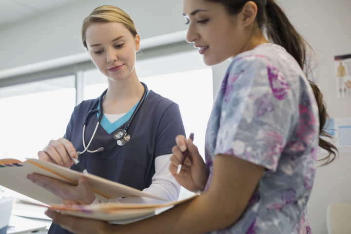 Two health care professionals look over medical files. One wears a dark blue scrub top with a stethoscope. The other person wears a scrub top with a colorful print.